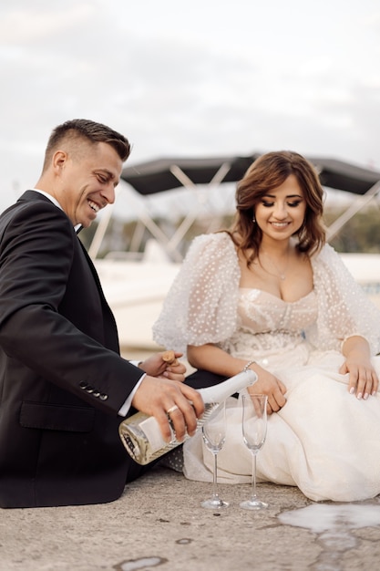Photo de jeune couple, mariée et marié en robes de mariée sont assis sur un pont en béton avec des verres de vin et de champagne. passer du temps ensemble, heureux.