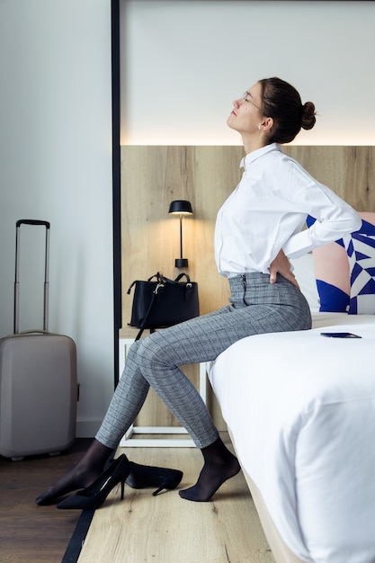 Photo d'une jeune femme d'affaires fatiguée souffrant de maux de dos assise sur le lit dans une chambre d'hôtel.