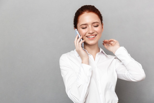 Photo d'une jeune femme d'affaires heureuse et jolie rousse posant isolée sur un mur gris parlant par téléphone portable.