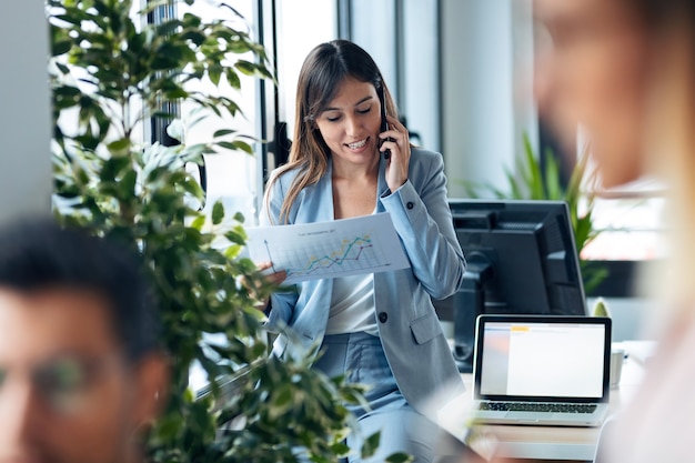 Photo d'une jeune femme d'affaires intelligente parlant avec un téléphone portable tout en examinant des documents dans un espace de travail moderne.