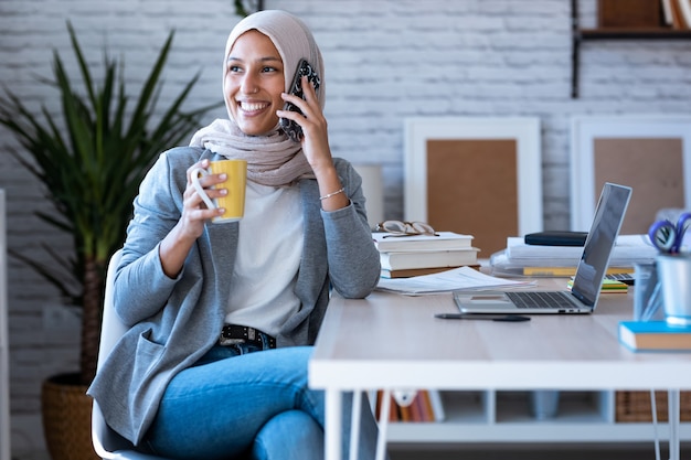 Photo d'une jeune femme d'affaires musulmane souriante portant le hijab parlant avec un téléphone portable assis dans le bureau.