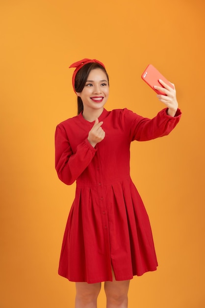 Photo d'une jeune femme asiatique heureuse prenant un selfie avec son téléphone portable tout en se tenant isolée sur fond orange