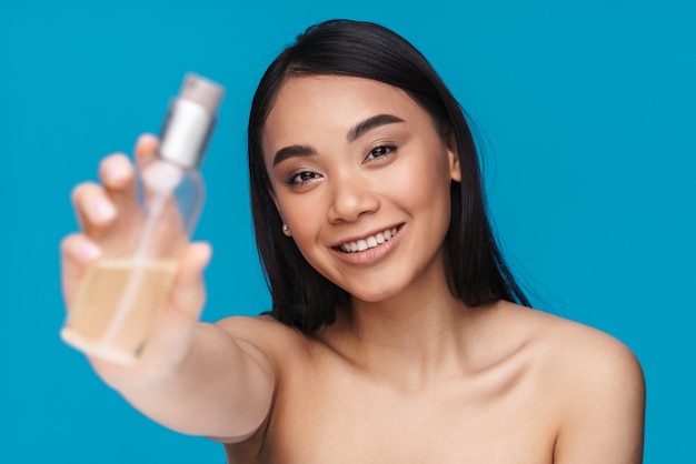 Photo d'une jeune femme asiatique joyeuse posant isolée sur un mur bleu montrant une bouteille d'huile cosmétique.