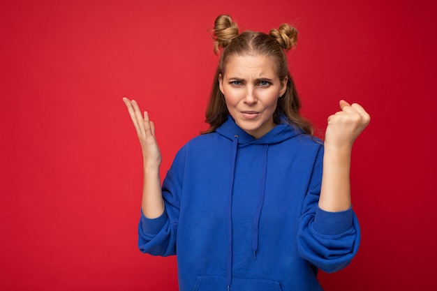 Photo d'une jeune femme blonde séduisante agressive émotionnelle avec deux cornes avec des émotions sincères portant un sweat à capuche bleu vif isolé sur fond rouge avec un espace libre et montrant le poing.