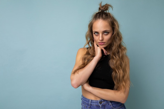 Photo de jeune femme blonde séduisante mode européenne portant un t-shirt haut noir isolé sur un mur bleu avec copie espace avec une expression sérieuse sur le visage. Personne de sexe féminin simple et naturelle.