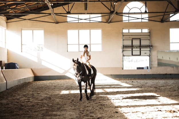 Photo d'une jeune femme blonde sérieuse et concentrée portant un chapeau à cheval dans la campagne à corral sur la zone d'entraînement.