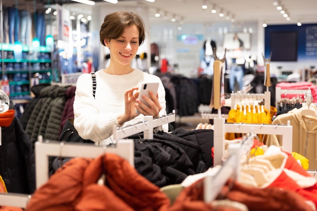 Photo d'une jeune femme brune séduisante avec une coupe de cheveux courte dans un pull blanc choisit des vêtements élégants et décontractés dans un magasin d'un centre commercial à l'aide d'un téléphone portable