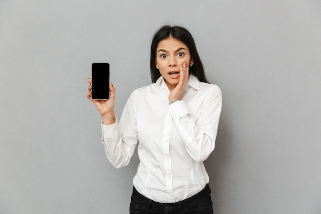 Photo de jeune femme caucasienne de 30 ans en tenue de soirée démontrant un écran noir de smartphone et exprimant la surprise, isolé sur fond gris