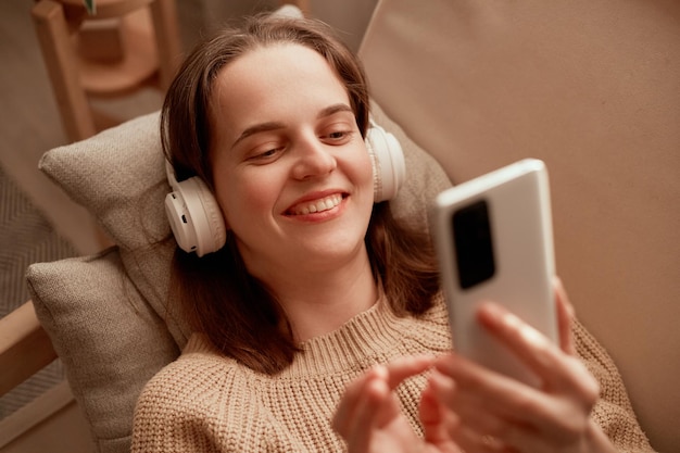 Photo d'une jeune femme caucasienne adulte portant des écouteurs tenant un téléphone portable allongée sur un canapé dans son appartement à l'aide d'un téléphone portable pour vérifier les réseaux sociaux ou discuter entre les mains