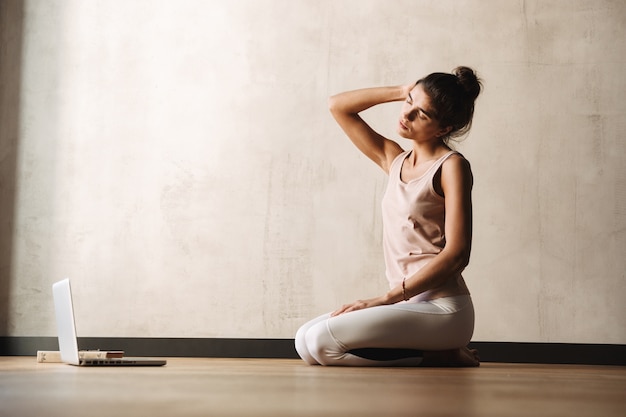 Photo d'une jeune femme concentrée en tenue de sport faisant des exercices les yeux fermés et utilisant un ordinateur portable assis par terre à la maison