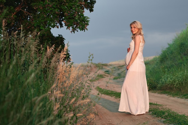 Photo de jeune femme enceinte en élégante robe blanche élégante posant dans le parc contre le ciel sombre. Bannière horizontale