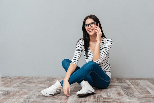 Photo d'une jeune femme heureuse portant des lunettes assise sur le sol tout en parlant par téléphone sur une surface grise. Regarder de côté.