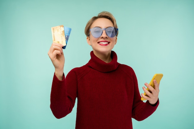 Photo de jeune femme heureuse posant isolé sur fond de mur bleu à l'aide de téléphone mobile tenant une carte de débit.