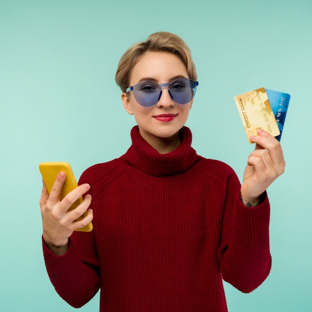 Photo de jeune femme heureuse posant isolé sur fond de mur bleu à l'aide de téléphone mobile tenant une carte de débit.
