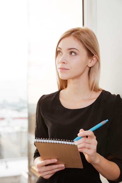 Photo d'une jeune femme joyeuse debout au bureau tenant un cahier tout en écrivant des notes avec un stylo. Regarder de côté.