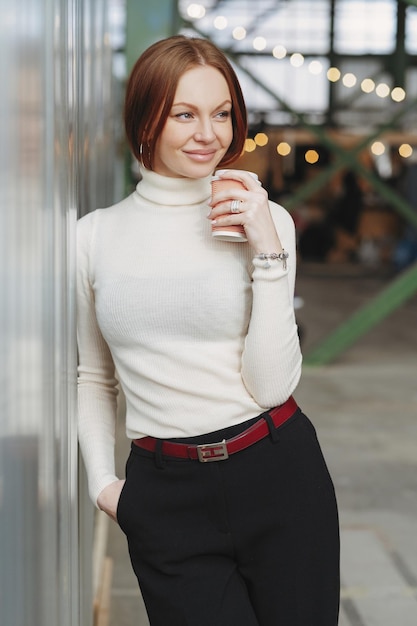 Photo d'une jeune femme joyeuse porte des vêtements élégants vêtus d'un pull blanc et d'un pantalon noir boit une tasse de café jetable rêve de quelque chose d'agréable garde le regard de côté Concept de repos