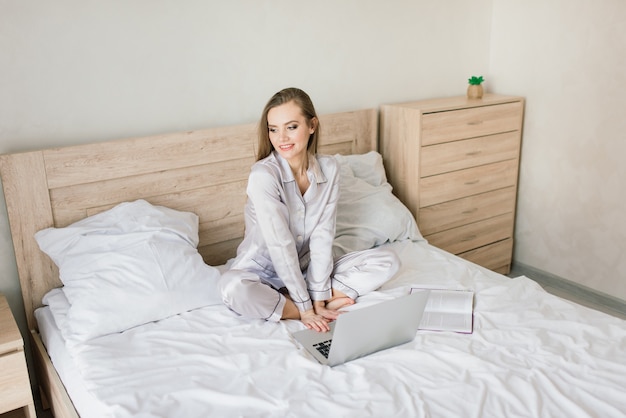 Photo de jeune femme joyeuse en pyjama en tapant sur un ordinateur portable et souriant alors qu'il était assis sur le lit dans la chambre