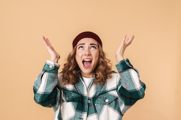 Photo de jeune femme nerveuse en tricot chapeau criant et regardant vers le haut isolé sur beige