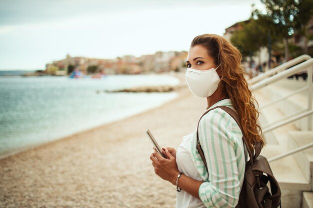 Photo d'une jeune femme portant un masque de protection N95 et utilisant un smartphone tout en profitant de la plage pendant le COVID-19.