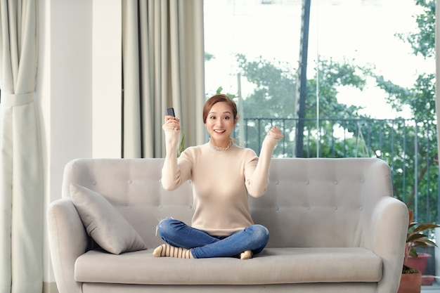 Photo d'une jeune femme positivement surprise sur un canapé à l'intérieur à la maison regarder la télévision faire un geste gagnant.