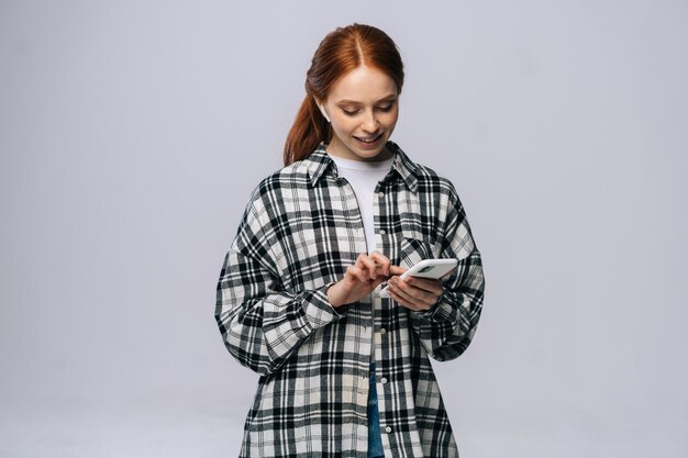 Photo d'une jeune femme rousse positive utilisant des écouteurs de technologie moderne écoutez une chanson populaire en choisissant le téléphone suivant la piste audio sur fond blanc isolé