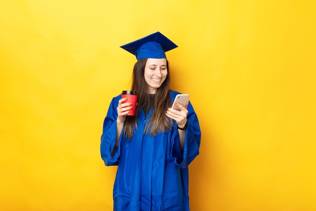 Photo de jeune femme souriante diplômée et à l'aide de smartphone et tenant une tasse de café