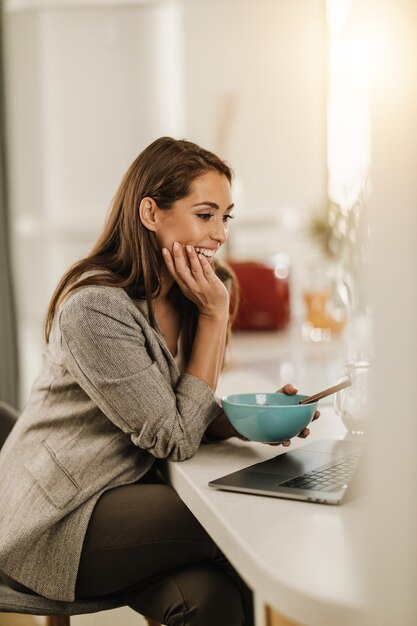 Photo d'une jeune femme souriante utilisant son ordinateur portable tout en prenant son petit-déjeuner à la maison.