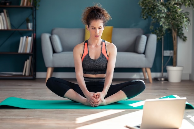 Photo d'une jeune femme sportive confiante faisant des exercices hypopressifs après des cours de gym en ligne via un ordinateur portable au sol dans son salon à la maison.