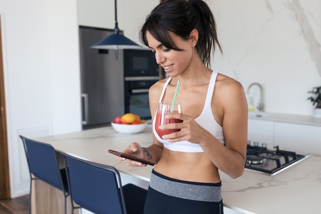 Photo d'une jeune femme sportive utilisant son téléphone portable tout en buvant un smoothie aux fraises dans la cuisine à la maison.