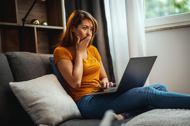 Photo d'une jeune femme surprise assise sur le canapé et utilisant son ordinateur portable à la maison.