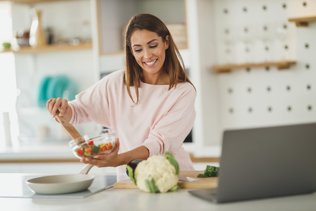 Photo d'une jeune femme utilisant un ordinateur portable pour créer un blog vidéo avec quelqu'un tout en préparant un repas sain à la maison.