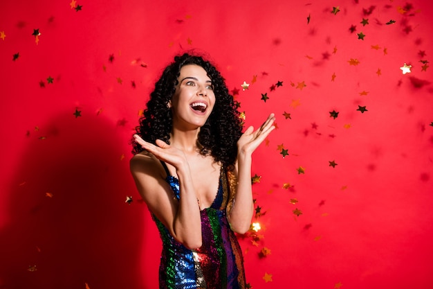 Photo d'une jeune fille étonnée, bouche ouverte, regarde l'espace vide, les étoiles dorées tombent porter une mini robe brillante isolée sur fond de couleur rouge vif