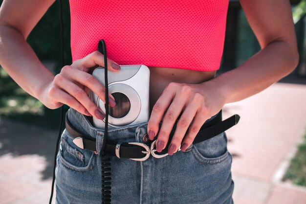 Une photo de jeune fille mettant le lecteur de musique en pantalon jeans. Le lecteur de musique est blanc et connecté aux écouteurs par un cordon.