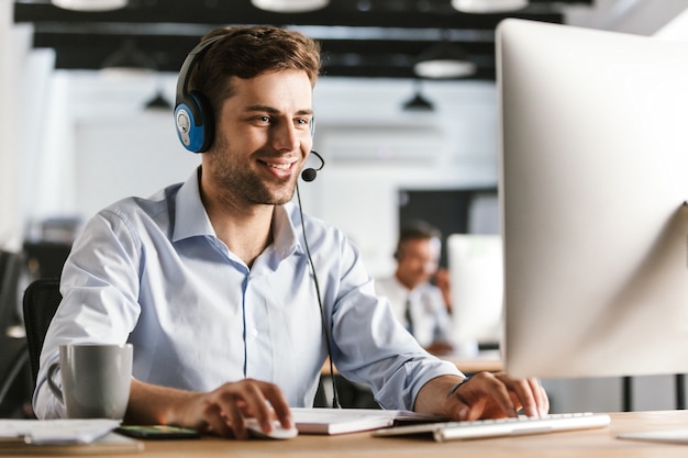 Photo photo d'un jeune homme de 20 ans portant des vêtements de bureau et un casque, souriant et parlant avec des clients dans un centre d'appels