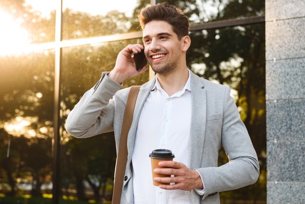 Photo de jeune homme d'affaires en costume, parler au téléphone mobile, debout en plein air près du bâtiment avec du café à emporter