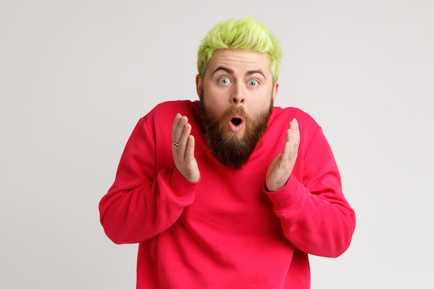 Photo d'un jeune homme barbu choqué étonné aux cheveux jaunes regarde la caméra avec la bouche ouverte levant les paumes impressionné par des nouvelles choquantes Prise de vue en studio intérieur isolée sur fond gris