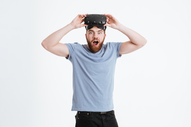 Photo d'un jeune homme barbu choqué portant un appareil de réalité virtuelle debout et regardant à l'avant.