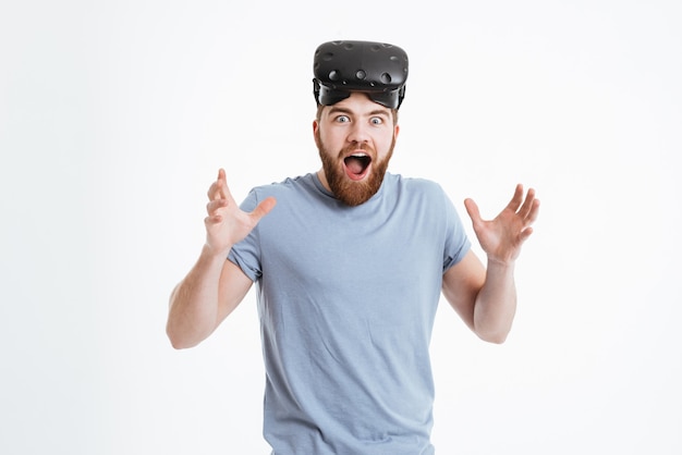 Photo d'un jeune homme barbu choqué portant un appareil de réalité virtuelle debout et regardant de face.