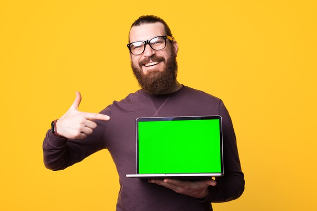 Photo d'un jeune homme barbu tenant un ordinateur sourit et le pointe du doigt