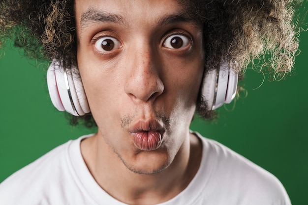 Photo photo d'un jeune homme bouclé surpris posant isolé sur un mur vert écoutant de la musique avec des écouteurs.