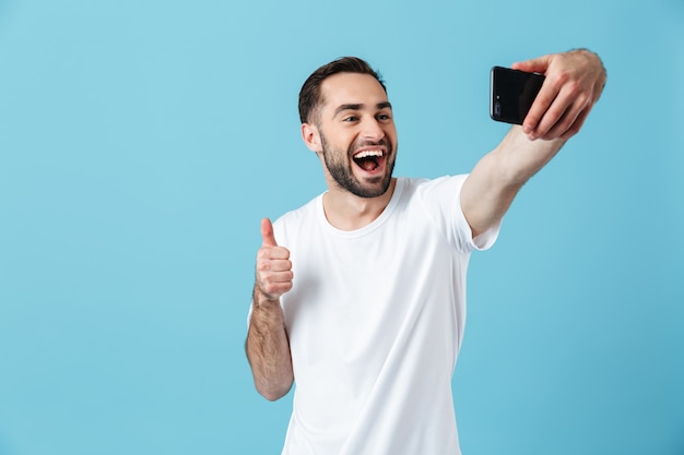 Photo de jeune homme brune portant un t-shirt basique riant et prenant selfie sur smartphone isolé sur bleu