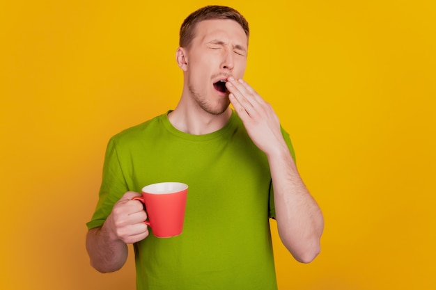 Photo de jeune homme endormi tôt le matin bâillement boire une tasse de café chaud isolé sur fond de couleur jaune