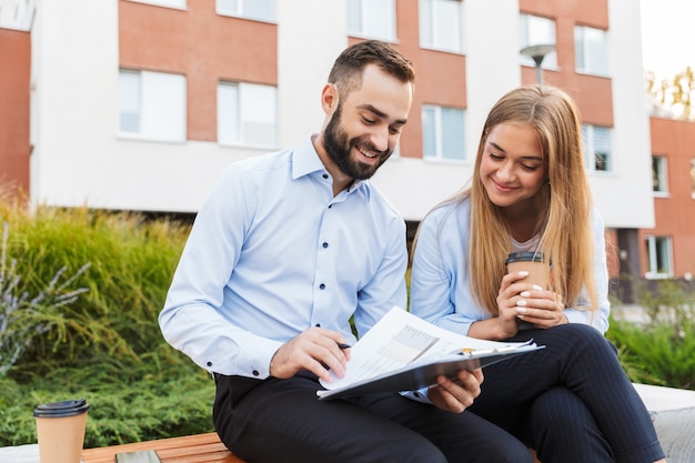 Photo d'un jeune homme et femme joyeux et joyeux, des hommes d'affaires à l'extérieur dans la rue tenant un presse-papiers avec des plans d'affaires de documents parlant entre eux en buvant du café.