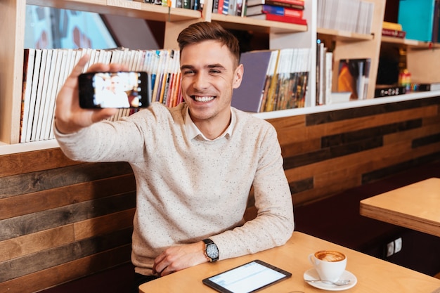 Photo d'un jeune homme gai vêtu d'une chemise assis dans un café et faisant un selfie par son téléphone.