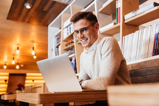 Photo d'un jeune homme heureux séduisant portant des lunettes assis dans un café tout en utilisant un ordinateur portable.