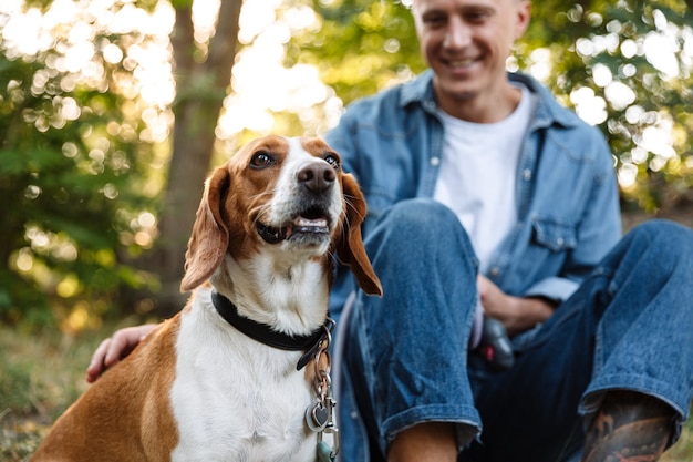 Photo d'un jeune homme heureux en vêtements en jean assis sur le sol dans un parc avec son chien canin