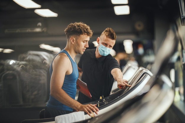 Photo d'un jeune homme musclé marchant sur le tapis roulant de la salle de sport. Il travaille avec un entraîneur personnel.