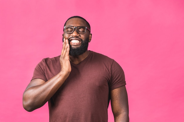 Photo d'un jeune homme noir afro-américain isolé sur fond rose souffrant d'une terrible douleur aux dents, la montrant sur son visage avec éclat, l'air troublé et affligé.