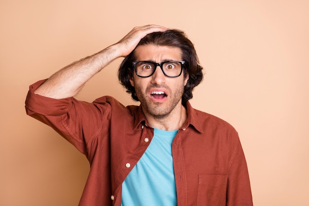 Photo de jeune homme perplexe main tête bouche ouverte look appareil photo porter des spécifications t-shirt marron isolé sur fond de couleur beige