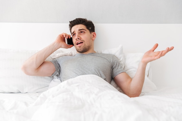 Photo de jeune homme de race blanche couché seul dans son lit avec du linge blanc et ayant un appel mobile important